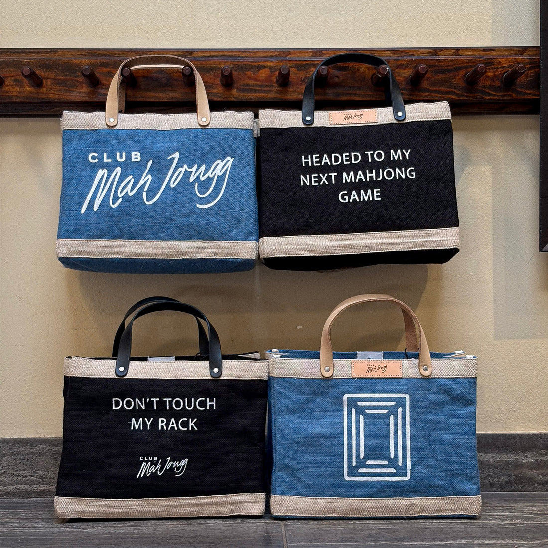 Four canvas tote bags arranged hanging from wooden pegs. The bags feature blue and black jute bags with beige trim, leather handles, and white printed text or graphics.