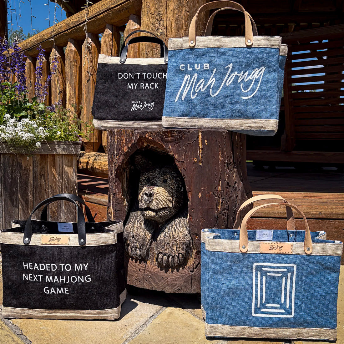 Four jute tote bags with leather handles displayed outdoors in front of a carved wooden bear and log railin. The bags feature different printed designs: one black with white text reading &
