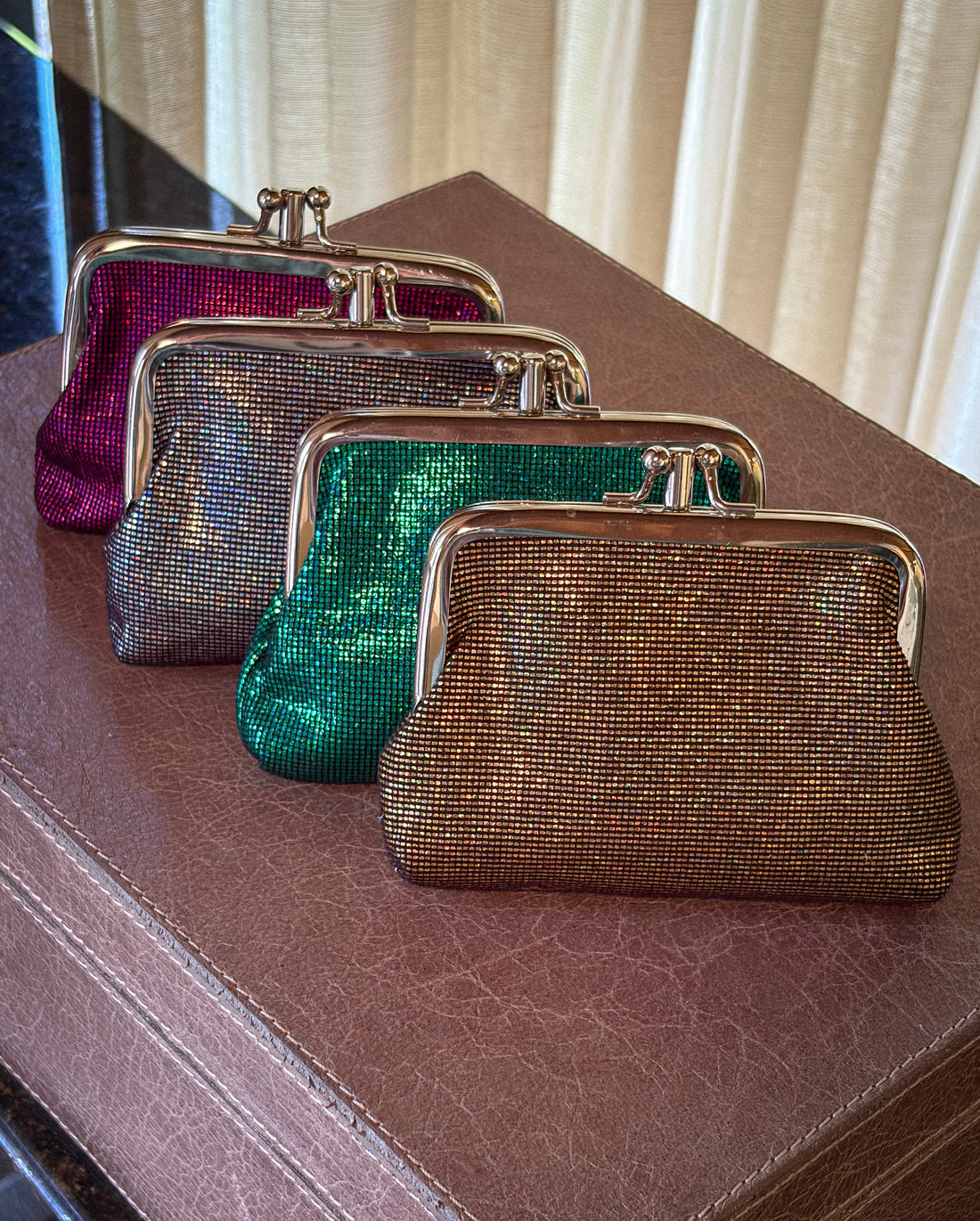 Four small kiss-lock clutch coin purse bags with metallic mesh bodies are arranged diagonally on a brown textured surface. The front bag is gold, the next is green, then silver, and last is magenta.