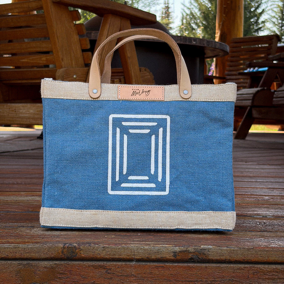 A blue Club Mah Jongg jute Apolis bag with Club Mah Jongg patch and icon -- white geometric line-art design. The bag is photographed outdoors on a wooden deck with wooden chairs and trees in the background.