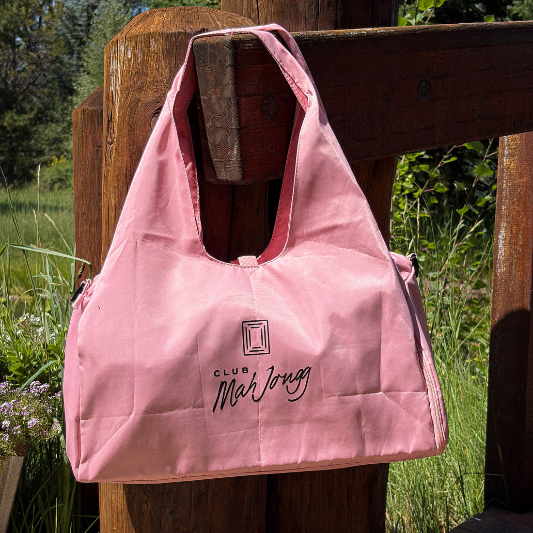 A pink Club Mah Jongg nylon bag with black logo text, hanging on a rustic wooden fence post in an outdoor garden setting.