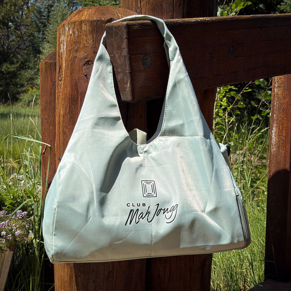 A light sage green Club Mah Jongg nylon zippered back with black logo text, hanging from a rustic wooden fence post in an outdoor grassy setting.