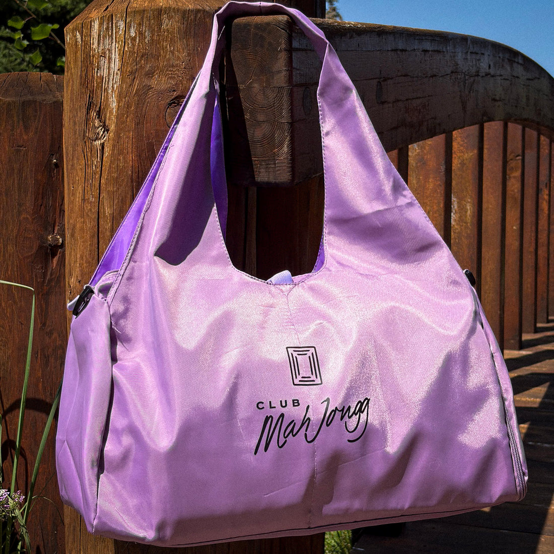 A lavender-purple Club Mah Jongg nylon tote bag hanging from a wooden fence outdoors. The bag features the &