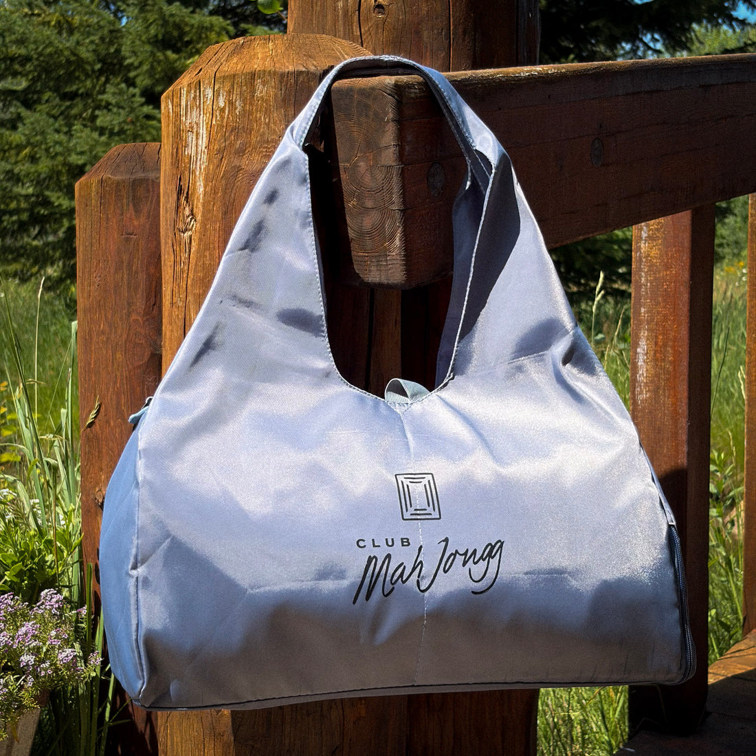 A light blue-gray Club Mahjongg zippered bag with shoulder straps, hanging from a bridges wooden railing with green foliage in the background.