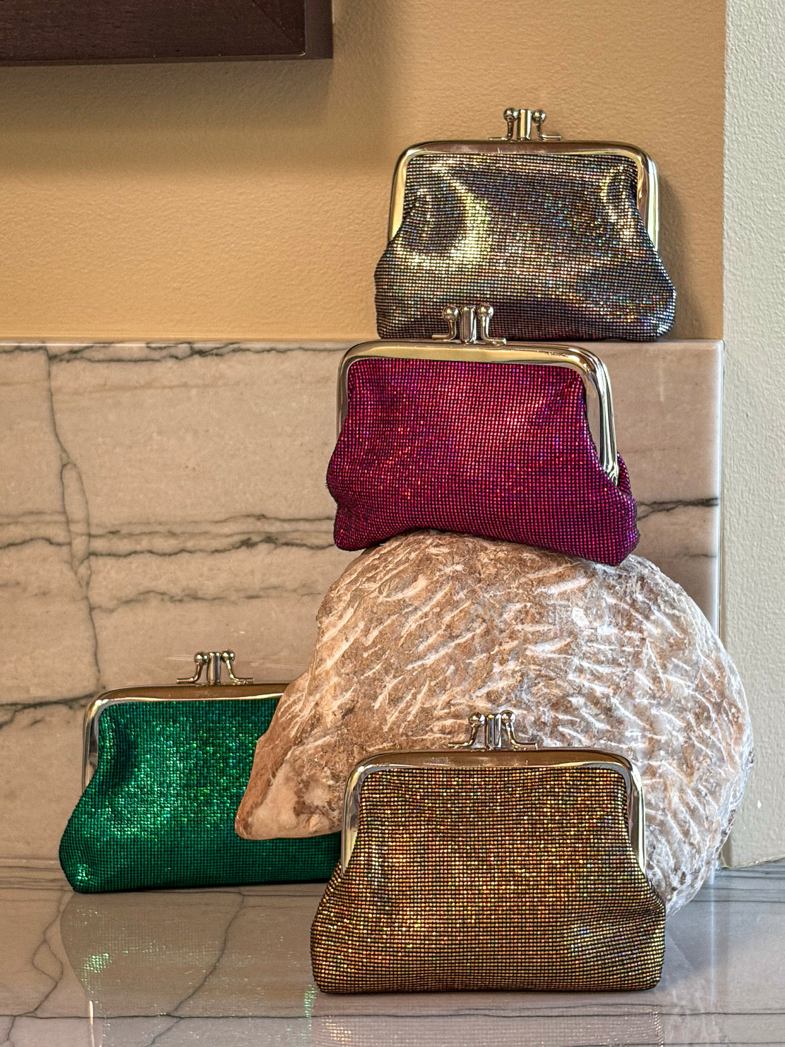Four sparkly kiss-lock clutch coin purses in silver, magenta, green, and gold, stacked and arranged on a marble surface against a beige wall.
