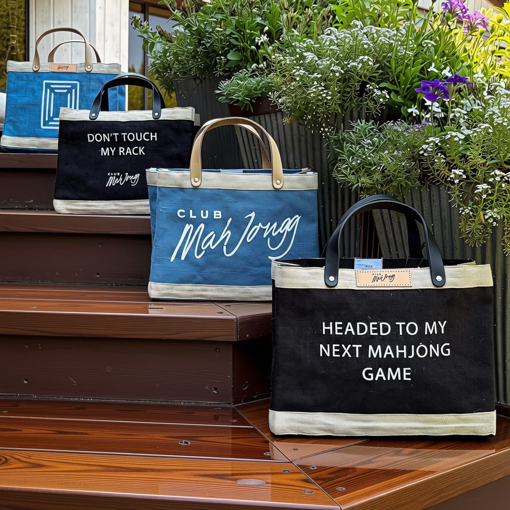 Four jute tote bags with leather handles arranged on wooden deck stairs next to potted flowers. The bags have different colors and printed phrases: one black bag says &