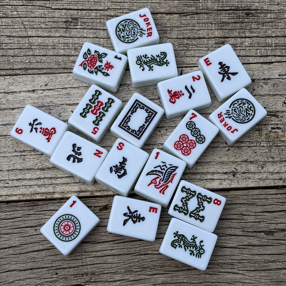 A set of classic white mahjong tiles from Club Mah Jongg with black, red, and green traditional Chinese characters and symbols, scattered on a rustic wooden surface. 