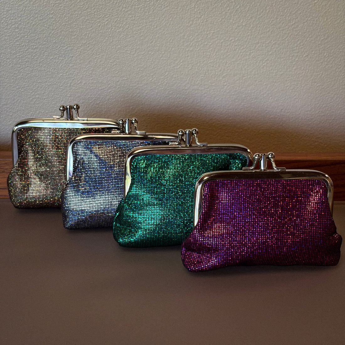 Four small kiss-lock clutch bags with iridescent glitter finishes lined up on a brown surface against a beige wall. The bags show a color-shifting effect with different hues including silver, blue, green, and purple.